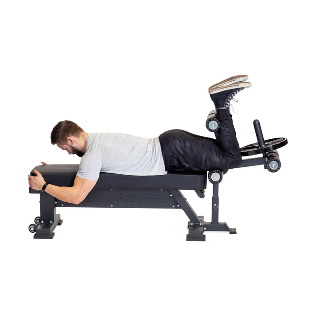A man in athletic wear demonstrates a hamstring curl by lying face down and curling his legs with weights, using Bells of Steel Bench Attachments.