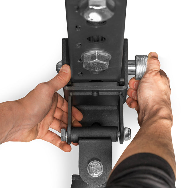 Close-up of hands adjusting a Bells of Steel Landmine Rack Attachment, featuring a black metal piece with a silver knob and UHMW-lined sleeve. The white background emphasizes its detailed components and bolts, perfect for enhancing strength workouts.