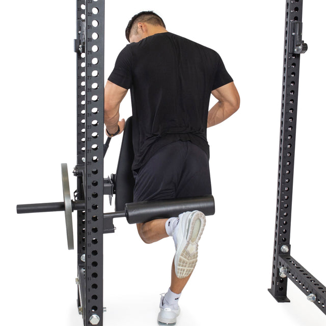 A man in black athletic wear uses the Bells of Steel Leg Curl / Leg Extension Rack Attachment, gripping the handles for support while performing a sissy squat. The plain white background highlights the equipment.