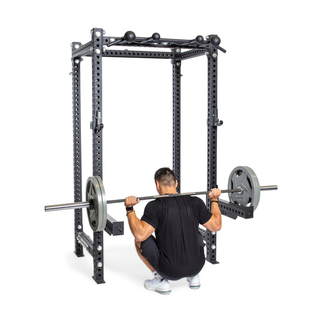 A man in athletic wear performs a barbell squat on the Bells of Steel Manticore Four Post Power Rack - Prebuilt (3" x 3", 1" Holes), holding a loaded barbell across his shoulders—an ideal addition to any home gym.