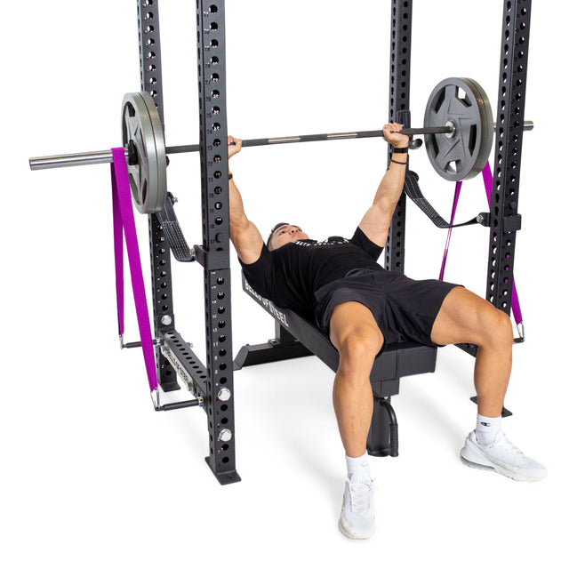 A person bench presses with a barbell and resistance bands on the Bells of Steel Manticore Four Post Power Rack (Prebuilt, 3" x 3", 1" Holes), wearing athletic clothing and white sneakers in their home gym.