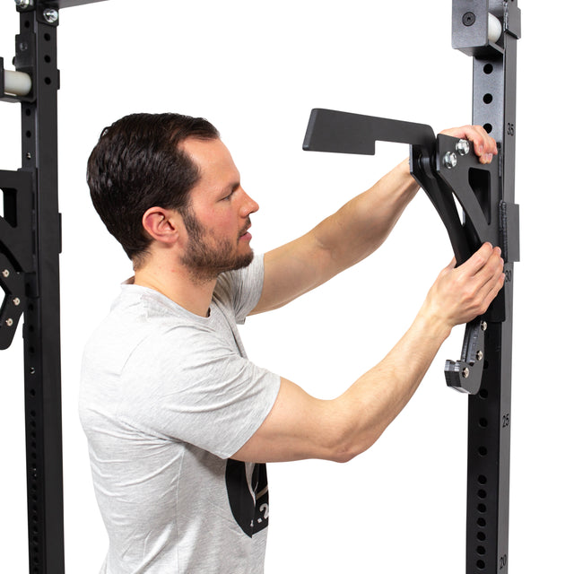 A man in a light gray t-shirt adjusts a black Bells of Steel Monolift Rack Attachment with UHMW plastic to a squat rack, highlighting its space-saving design against a white background.
