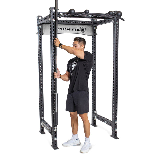 A man in athletic wear adjusts a barbell inside a black Bells of Steel power rack with multiple holes and pull-up attachments. A Bells of Steel Vertical Mount Barbell Holder Rack Attachment is installed for added convenience.