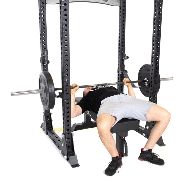 A person in a black shirt and gray shorts is bench pressing with a barbell inside the Bells of Steel Manticore Flat Foot Power Rack - Prebuilt (3" x 3", 1" Holes) in a home gym with a white background.