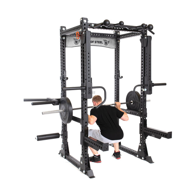 A man performs a barbell squat inside the Bells of Steel Manticore Flat Foot Power Rack - Prebuilt (3" x 3", 1" Holes), equipped with weights and attachments, perfect for home gyms, shown against a plain white background.