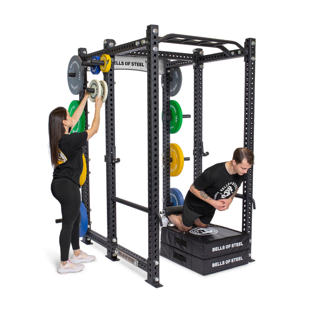 A woman adjusts weight plates on a Bells of Steel Manticore Six Post Power Rack, demonstrating its modular design, while a man exercises on a pad nearby. Both are dressed in workout attire.