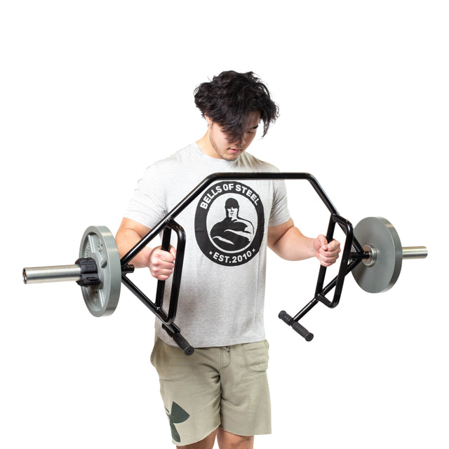 A person wearing a gray Bells of Steel t-shirt and khaki shorts holds the Bells of Steel Open Trap Bar / Hex Bar, looking down, against a plain white background.