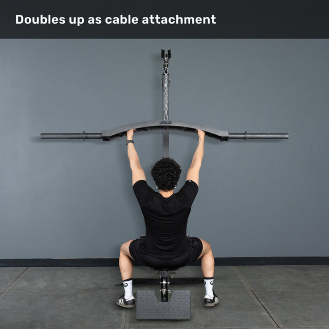 A person sits on a bench facing a wall, holding the Bells of Steel Arch Nemesis Swiss Bar attached to an overhead cable machine, with the caption "Doubles up as cable attachment" above.