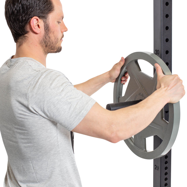 A man in a light gray t-shirt loads a large plate onto Bells of Steel Pin Plate Pegs, demonstrating convenient Olympic weight plate storage on rack-mountable pegs in the gym against a white background.