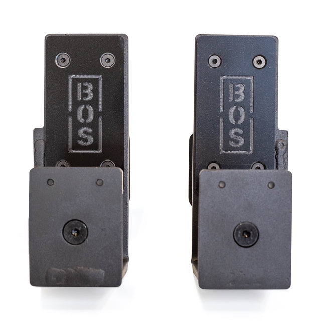 Two black metal Roller J-Cups by Bells of Steel, each engraved with "BOS," feature multiple screws and square bases. Designed to protect barbells during intense bench presses, they are shown against a white background.