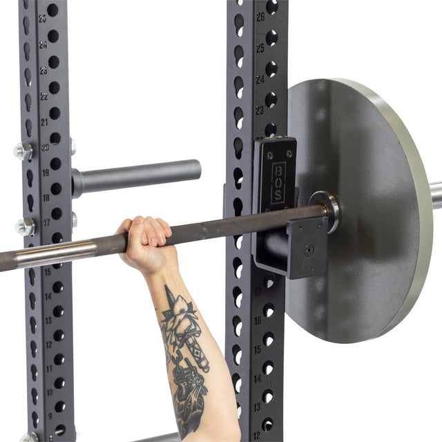 A tattooed arm grips a barbell loaded with weight plates on a squat rack equipped with Bells of Steel Roller J-Cups, preparing for a lift against a white background.