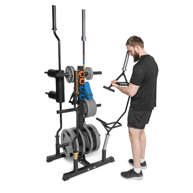 A man in a black athletic outfit stands next to the Bells of Steel Residential Bumper Plate Tree, efficiently organizing weight plates. Among his home gym equipment, he lifts a metal attachment with determination.