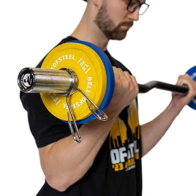 Someone in glasses and a black T-shirt performs a bicep curl with an Olympic barbell, which features polished chrome sleeves and yellow and blue weight plates secured by Bells of Steel Spring Collars. The person's face is not visible.