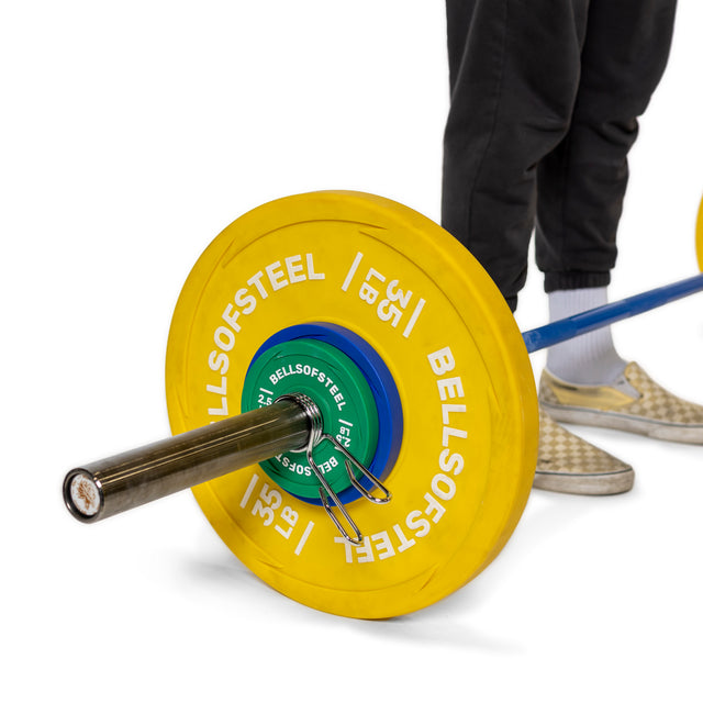 A person in black pants and checkered slip-ons admires a barbell with yellow Bells of Steel weight plates, secured by Spring Collars on its shiny chrome Olympic sleeves, set against a pristine white background.