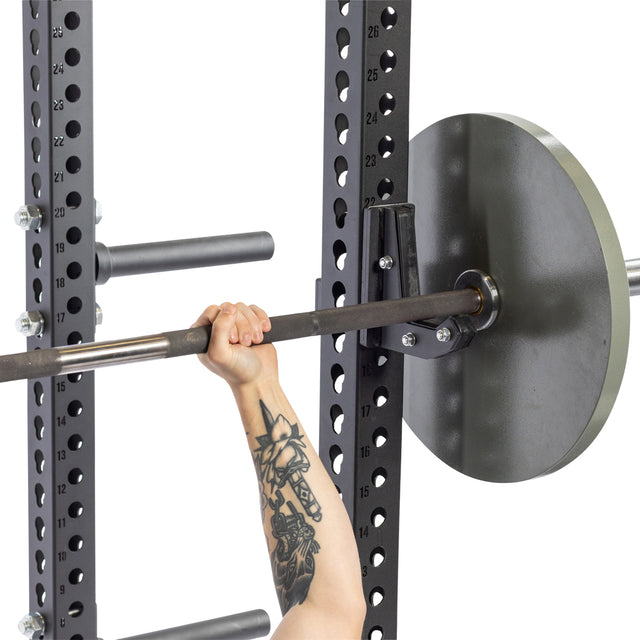 A tattooed lifter grips a barbell on a squat rack equipped with Bells of Steel Sandwich J-Cups and storage pegs—essential accessories for serious lifters.