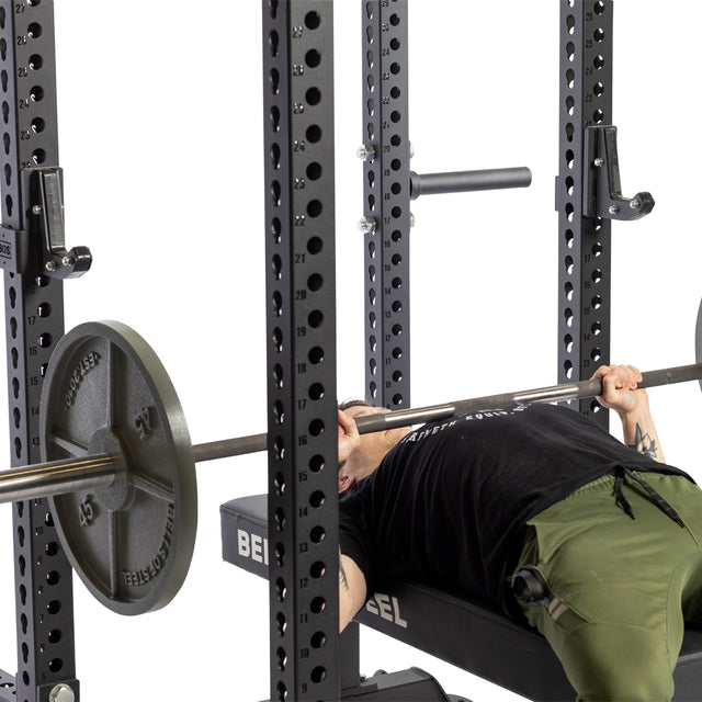 A person in green pants and a black shirt is lying on a bench, gripping a barbell with weight plates inside a power rack equipped with Bells of Steel Sandwich J-Cups, preparing to do a bench press.