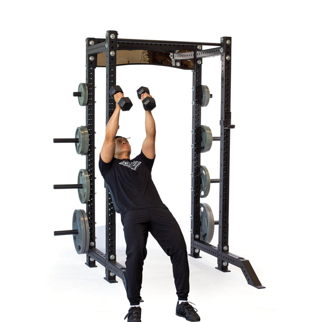 A person in black workout clothes performs a shoulder press with two dumbbells while leaning against the Bells of Steel Seal Row Pad Rack Attachment, which is mounted on a power rack loaded with weight plates, on a white background.
