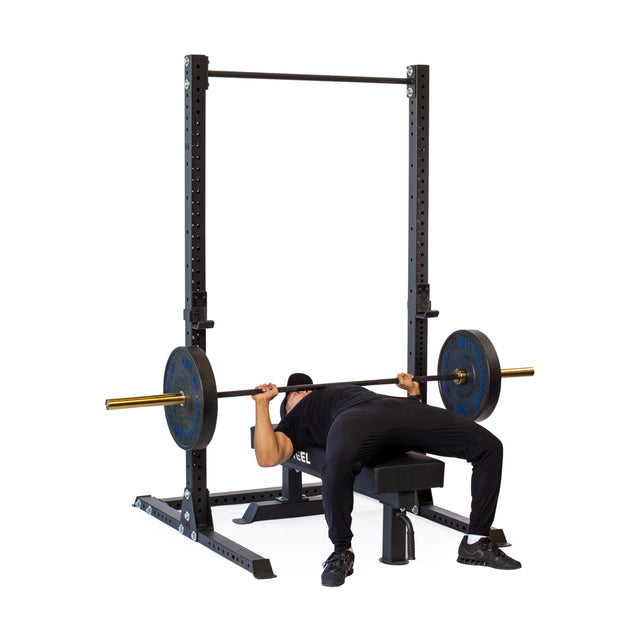 A person in black workout clothes bench presses with a loaded barbell on a bench under the Bells of Steel Hydra Squat Stand - Prebuilt (3" x 3", ⅝" Holes), isolated on a white background.