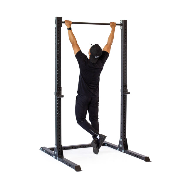 A person in black athletic wear and a cap does a pull-up on the Bells of Steel Hydra Squat Stand - Prebuilt (3" x 3", ⅝" Holes), facing away from the camera against a white background.