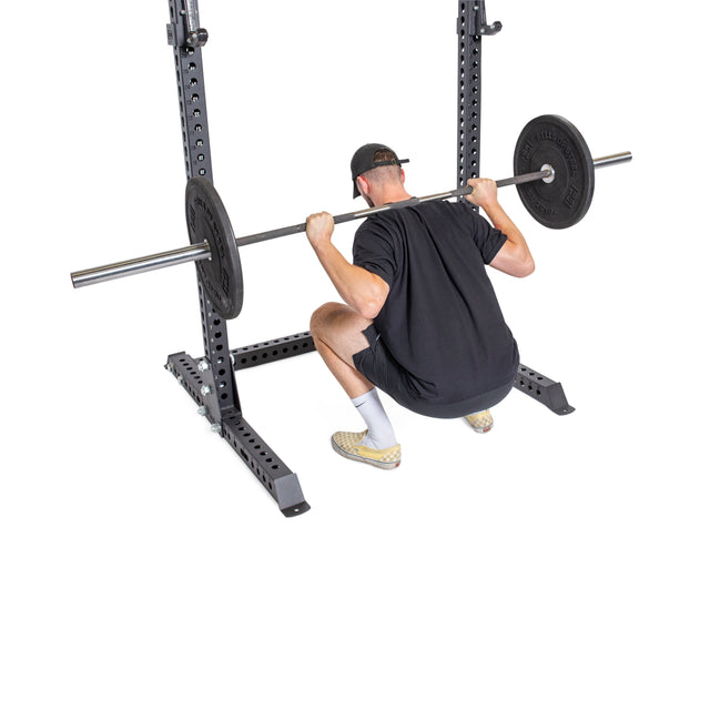 A man in black workout gear squats with a barbell inside the Bells of Steel Manticore Squat Stand Builder (3" x 3", 1" Holes), viewed from behind on a white background—ideal for any home gym.