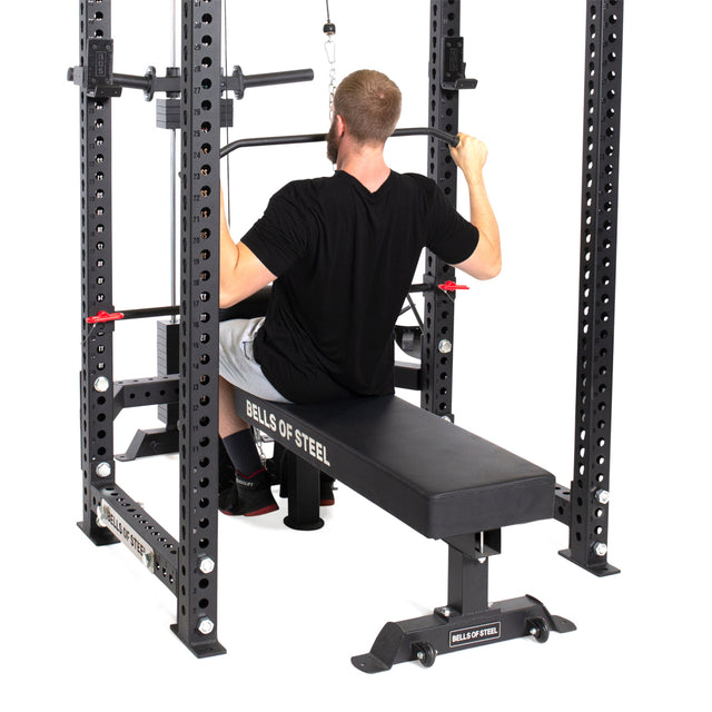 A man in a black shirt and gray shorts uses the Bells of Steel Lat Pulldown & Low Row Rack Attachment - Hydra & Manticore on a power rack with adjustable bench, pulling a wide bar with Olympic plates down toward his shoulders for cable exercises.