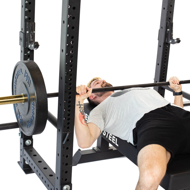 A man prepares for a bench press, lying on a bench inside a power rack with a barbell, using Bells of Steel Sandwich J-Cups for secure support.