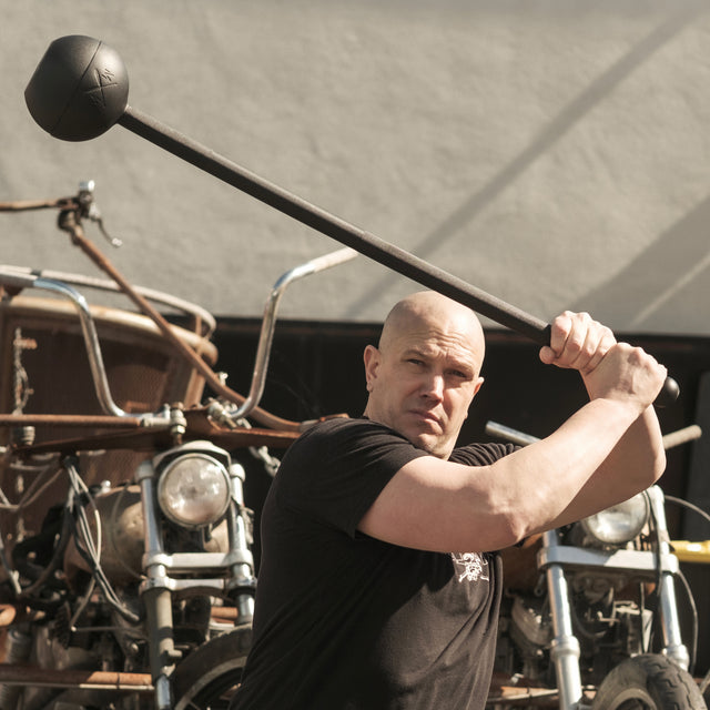 A person in a black shirt confidently holds a Bells of Steel Adjustable Macebell above their head, standing before vintage motorcycles. The blurred background highlights their core strength and the mace's dynamic presence.