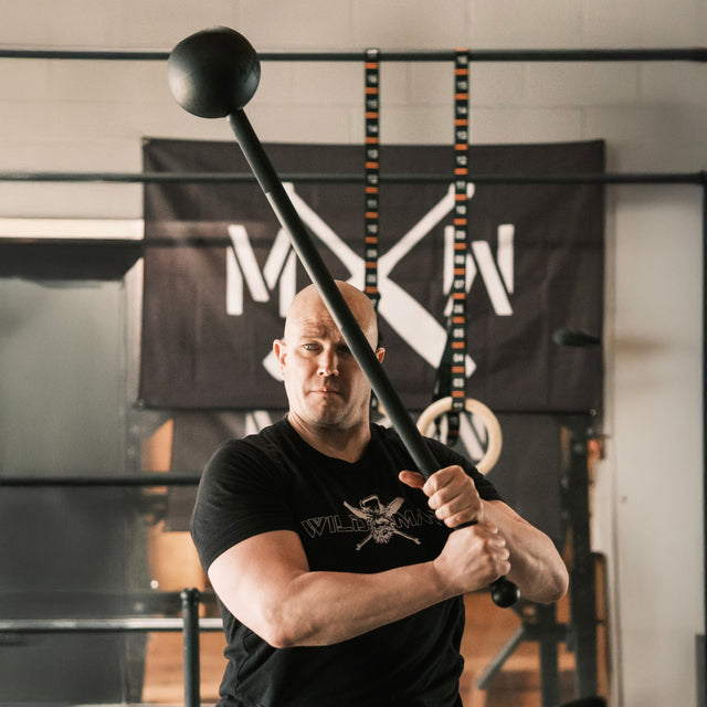 In the gym, a person in a black shirt skillfully swings the Bells of Steel Adjustable Macebell overhead, enhancing core strength and shoulder mobility. Surrounded by exercise equipment and inspiring wall banners, they create a motivating workout atmosphere.