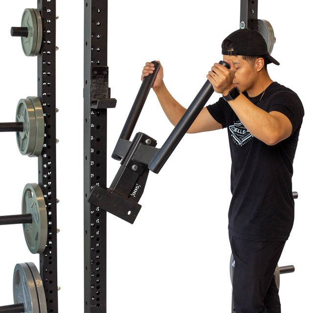 A person in a black shirt and cap adjusts the Bells of Steel Y Dip Bar Rack Attachment on the gym's heavy-duty rack, surrounded by weight plates. UHMW lining protects both the attachment and metal rack from wear.