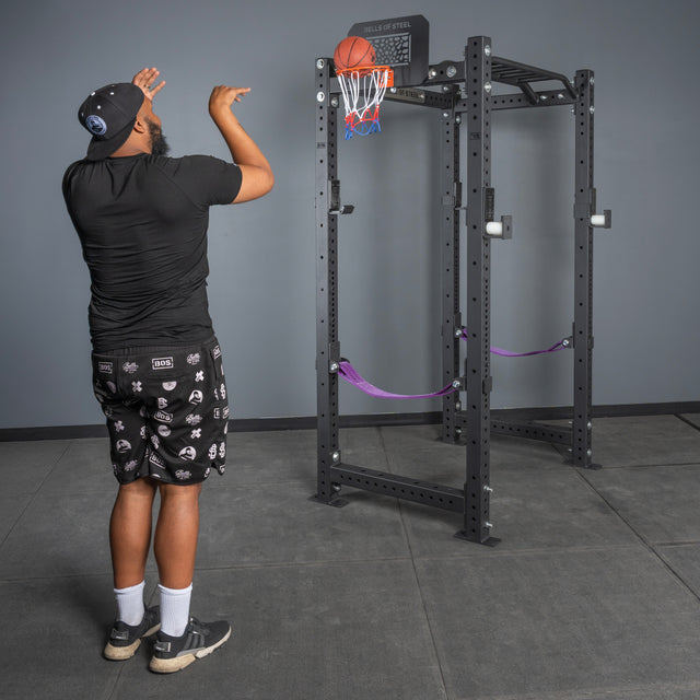 A person in a black shirt, patterned shorts, and a cap shoots the included ball at the Bells of Steel Rack Attached Basketball Hoop on a squat rack in a gym, showing how fun it is to add this hoop to your home gym.