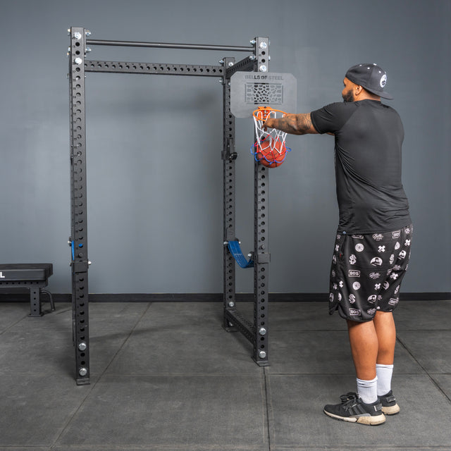 A man at the gym attaches resistance bands to a Bells of Steel Rack Attached Basketball Hoop w/ Ball, wearing a black shirt, patterned shorts, and a cap. The gym has gray walls and flooring.