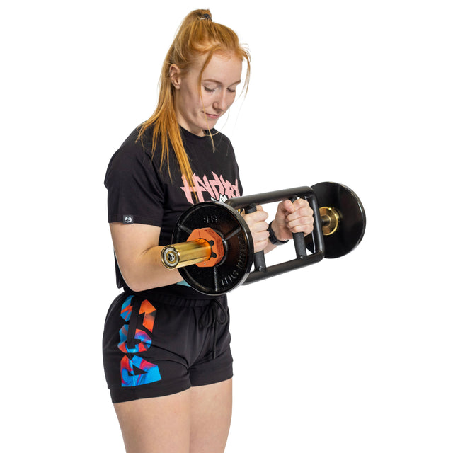 A woman with long red hair in a ponytail performs a bicep curl using a weight bar secured with Bells of Steel Magnetic Clamp Collars. She wears a black T-shirt and colorful shorts against a white background.