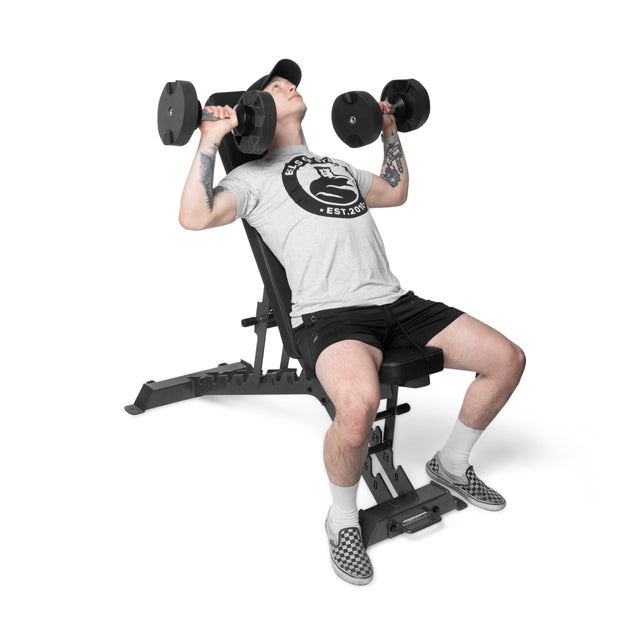 A person in a gray T-shirt, black shorts, and checkered slip-ons sits on the Bells of Steel Buzz-Saw Heavy-Duty Adjustable Bench, performing a shoulder press with dumbbells against a white background.