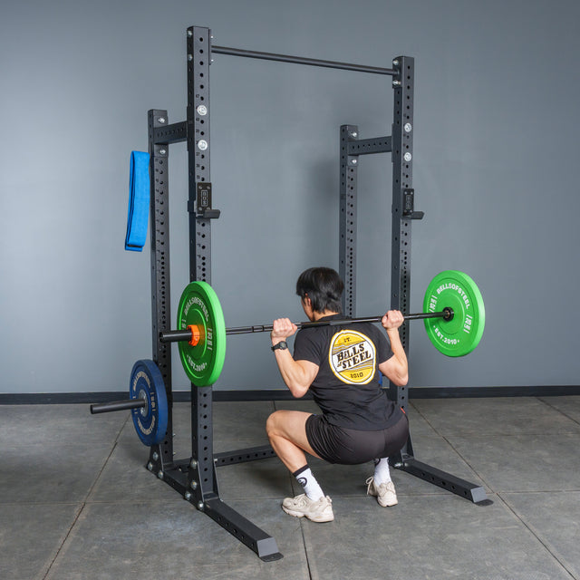 A person squats with a barbell loaded with green and blue plates on the Bells of Steel Hydra Half Rack Builder (3" x 3", ⅝" Holes) in a gym, facing away from the camera, wearing a black shirt, shorts, and white shoes.