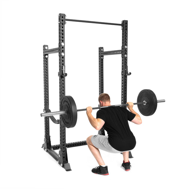 A person in a black shirt and gray shorts is performing a barbell squat inside the Bells of Steel Manticore Half Rack - Prebuilt (3" x 3", 1" Holes), perfect for any home gym, on a white background.