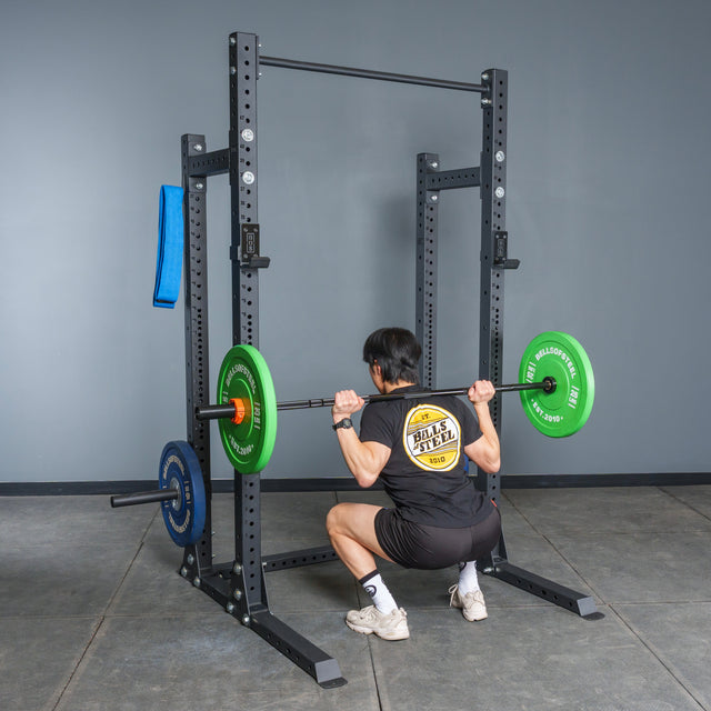 A person wearing a black shirt and shorts performs a barbell squat in a Bells of Steel Hydra Half Rack - Prebuilt (3" x 3", ⅝" Holes) in a gym, holding a barbell with green and blue plates across their upper back.