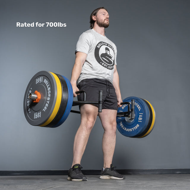 A man lifts the Bells of Steel Open Trap Bar / Hex Bar, rated for 700lbs, in a gym. He wears a grey t-shirt and black shorts. The bar features rotating sleeves; the background is a plain gray wall and floor.