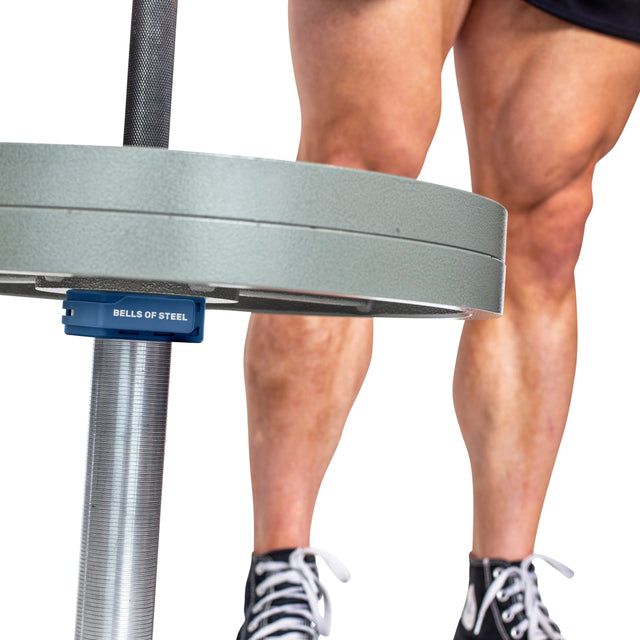Close-up of a person's muscular legs in black and white sneakers standing behind a loaded barbell, secured by Bells of Steel Magnetic Clamp Collars.