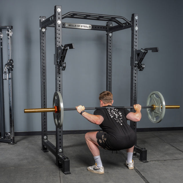 Wearing athletic clothing, a person performs a barbell squat in a Bells of Steel power rack equipped with the Monolift Rack Attachment, easily adjusting the barbell with weight plates on each side against gray gym walls.