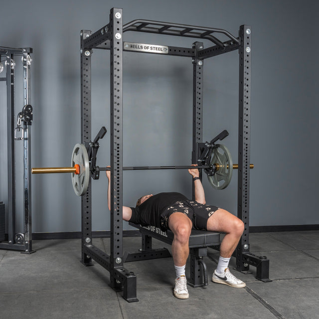 A person bench presses inside a Bells of Steel power rack, using the Monolift Rack Attachment with loaded weight plates. The black steel rack features a space-saving design and adjustable setup for efficient gym workouts.