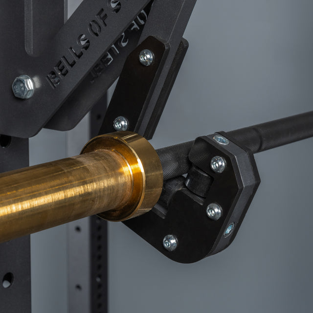 Close-up of a barbell with a golden sleeve secured in the Bells of Steel Monolift Rack Attachment, featuring UHMW plastic and a black metal holder fastened to gym equipment with bolts and washers for space-saving use.