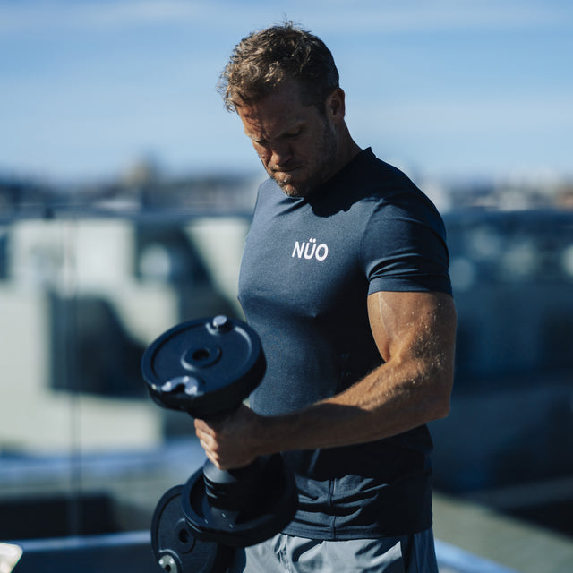A man lifts Bells of Steel NÜOBELL-S Adjustable Dumbbells outdoors on a sunny day, wearing a dark athletic shirt with blurred buildings and a blue sky in the background.