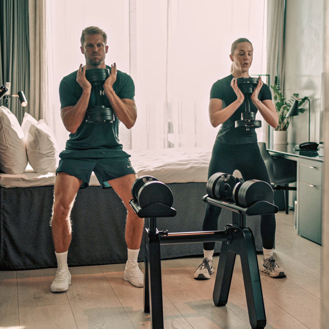 A man and woman work out in a bright home gym, each holding Bells of Steel NÜOBELL-S Adjustable Dumbbells at their chest while squatting next to a weight rack, with a bed and large window in the background.
