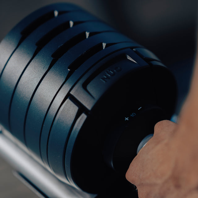 A hand grips the handle of a Bells of Steel NÜOBELL-S Adjustable Dumbbell, showing visible weight plates and a numbered selection dial—ideal for any home gym.