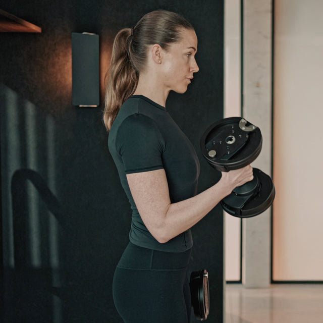 A woman with a ponytail in black athletic wear stands in her home gym, performing a bicep curl with Bells of Steel NÜOBELL-S Adjustable Dumbbells, facing sideways against a dark background with a wall sconce.