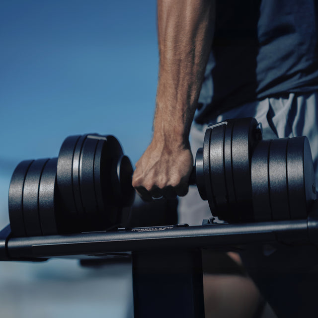 A close-up shows a hand gripping a Bells of Steel NÜOBELL-S Adjustable Dumbbell on the rack, with a muscular arm in athletic shorts, set against a blurred blue background in a modern home gym.