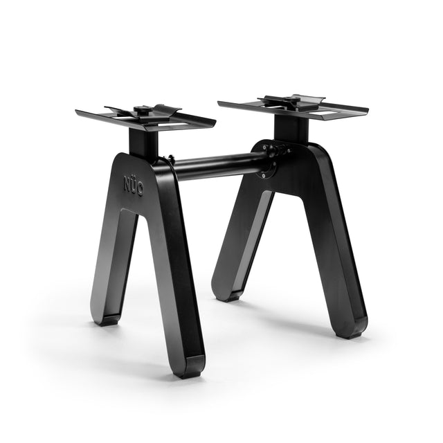 The NÜOBELL-S Floor Stand by Bells of Steel is a black, modern metal table base with two wide angled legs joined by a horizontal bar. "NÜO" is embossed on one leg. Designed to support a tabletop, it is photographed against a white background.