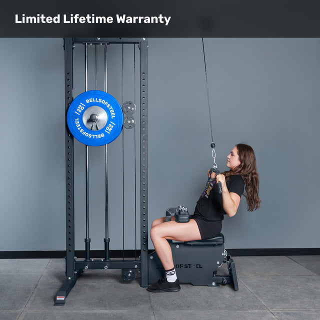 A woman uses the Bells of Steel Lat Pulldown Low Row Machine with blue weight plates and a selectorized stack in a gym. "Limited Lifetime Warranty" appears at the top.
