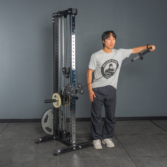 A person uses the Bells of Steel Cable Tower to perform a standing lateral raise in a gym with gray walls and flooring, highlighting effective cable machine exercises.