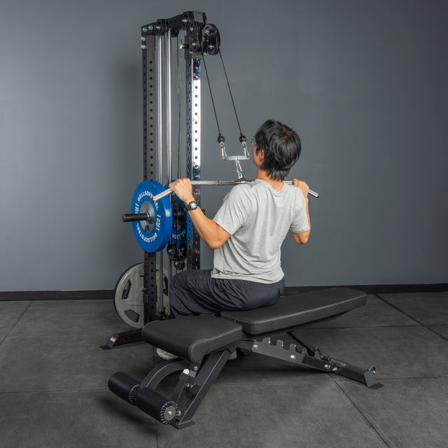 A person uses the Bells of Steel Cable Tower in the gym, seated on a workout bench and performing a wide grip lat pulldown with the weight stack.